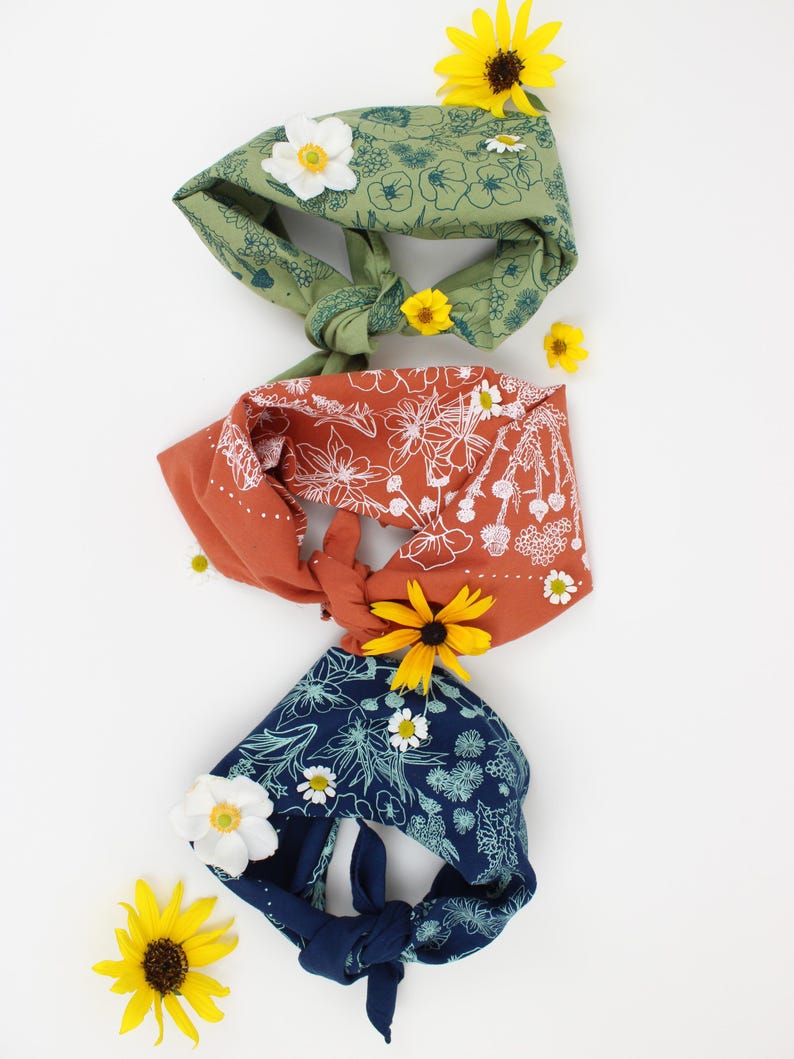Peut inclure: Trois bandanas en tissu avec des motifs floraux. Les bandanas sont nou&eacute;s et dispos&eacute;s verticalement. Le bandana du haut est vert avec des fleurs blanches, le bandana du milieu est orange avec des fleurs blanches et le bandana du bas est bleu avec des fleurs blanches. Il y a des fleurs jaunes et blanches &eacute;parpill&eacute;es autour des bandanas.