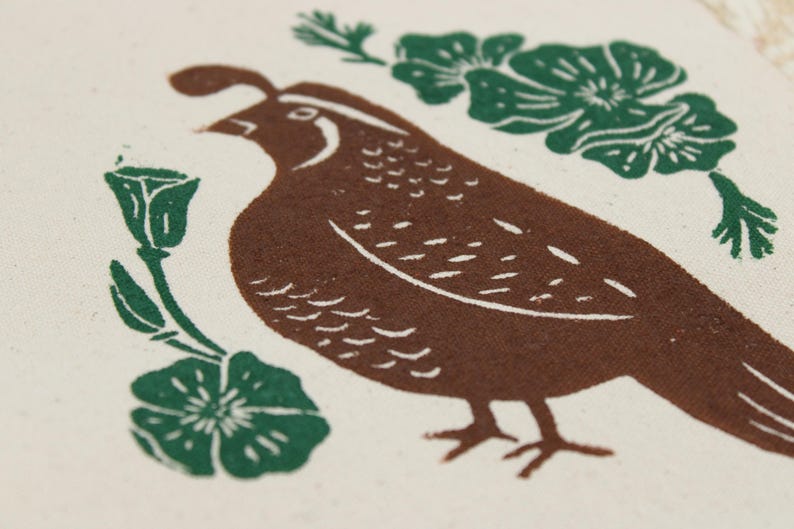 Op de afbeelding: Een cr&egrave;mekleurige stof met een bruine, in blokdruk uitgevoerde kwartel die naar links kijkt. De vogel is omringd door groene florale elementen, waaronder een kleine knop aan de linkerkant, een bloem linksonder en een grotere bloementros rechtsboven.