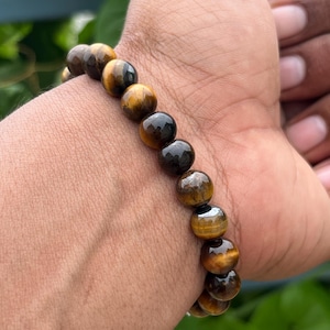 Può includere: Un braccialetto fatto di pietre di occhio di tigre lucide. Il braccialetto presenta una serie di perline rotonde, marroni e dorate infilate insieme. Il braccialetto è indossato su un polso.