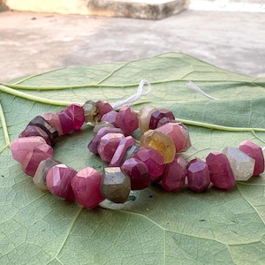 May include: A strand of faceted, irregular gemstone beads in shades of deep pink, magenta, translucent white, and muted olive green. The beads are strung on a thin white thread, showcasing a variety of natural shapes and semi-transparent textures.
