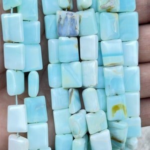 Puede incluir: Cadenas de cuentas rectangulares azul claro con un acabado pulido. Las cuentas están ensartadas, mostrando variaciones de color y patrones sutiles. La estética general es fresca y relajante, adecuada para la fabricación de joyas.