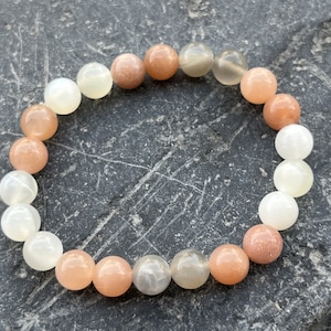 Könnte beinhalten: Ein Sonnensten-Armband mit weißen, pfirsichfarbenen und orangefarbenen Perlen. Das Armband besteht aus natürlichen Steinen und ist auf einem Gummiband gefädelt.