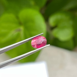 May include: A faceted, oval-shaped ruby gemstone held by tweezers. The ruby is a vibrant red color, with visible internal facets. The background features blurred green foliage and a white surface.