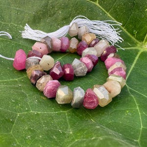 May include: A strand of faceted, multi-colored gemstone beads in shades of pink, magenta, tan, and grey. The beads are strung on a white thread with white tassels at the ends, arranged in a circular shape with irregular, polished surfaces.
