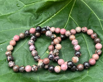 May include: Three beaded bracelets with pink, brown, and black stones rest on a large green leaf. The bracelets are made of round, polished beads, each approximately 0.8 cm in diameter. The beads have a speckled appearance.
