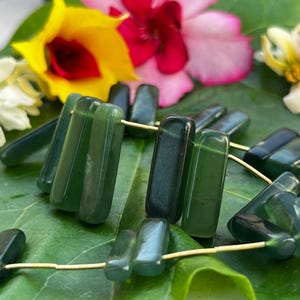 Könnte beinhalten: Dunkelgrüne, rechteckige Edelsteinperlen auf goldfarbenem Draht, präsentiert auf einem großen grünen Blatt. Der Hintergrund zeigt bunte Blumen in Gelb-, Rosa- und Rottönen.
