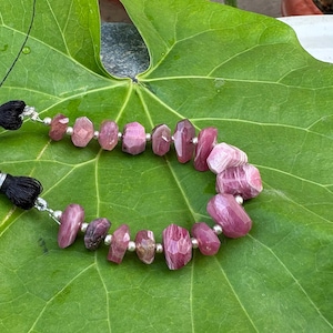 May include: A strand of faceted, irregular pink gemstone beads interspersed with small silver-toned metal spacers. The strand features two black thread tassels at the ends, with the beads arranged in a curved line showing varied shades of magenta and rose.