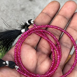 Peut inclure: Un collier de petites perles rondes rose vif, possiblement des pierres précieuses, est drapé sur une main. Les perles sont enfilées ensemble. Des pompons noirs avec des accents argentés sont visibles à l'extrémité du collier.