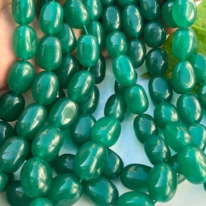 Puede incluir: Cadenas de cuentas verdes ovaladas y pulidas. Las cuentas están ensartadas, creando un collar o pulsera. Las cuentas tienen un acabado brillante, reflejando la luz y mostrando su color verde esmeralda intenso. El fondo es de un tono suave y neutro.