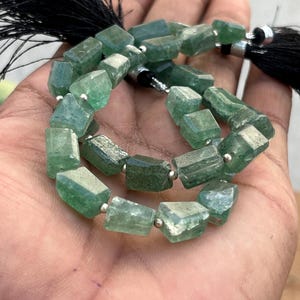 Peut inclure: Un fil de perles de pierres précieuses vertes de forme rectangulaire et facettée. Les perles sont enfilées avec des entretoises argentées et des pompons noirs à chaque extrémité. Les perles sont tenues dans une main.