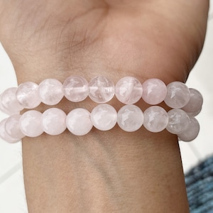 Pode incluir: Um bracelete de dupla corda feito de contas de quartzo rosa. As contas são redondas e lisas, e o bracelete é usado no pulso.