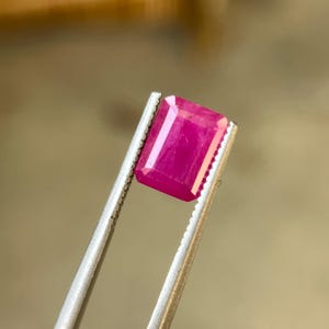 May include: A vibrant, rectangular-cut ruby gemstone held by silver tweezers. The ruby displays a rich, deep pink color with a polished surface. The gemstone's facets catch the light, showcasing its clarity and brilliance. A close-up view highlights the gem's quality.