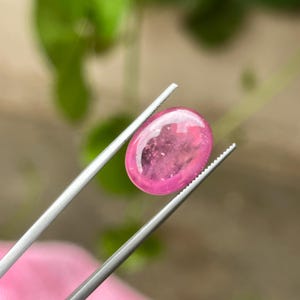 May include: A vibrant, oval-shaped pink gemstone with a smooth, polished cabochon surface. The translucent stone displays internal inclusions and is held securely by a pair of silver-toned metal tweezers with serrated tips for a firm grip.