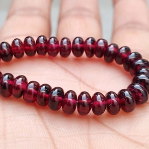 Puede incluir: Un brazalete hecho de cuentas de piedras preciosas de color rojo oscuro. Las cuentas son redondas y lisas, y están enhebradas en un cordón elástico transparente.