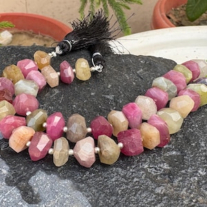 May include: A strand of faceted, multi-coloured tourmaline beads in shades of pink, yellow, and green, interspersed with small silver-toned metal spacer beads. The strand is finished with black thread tassels at both ends for a decorative look.