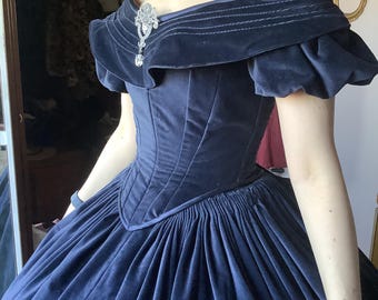 Puede incluir: Un vestido de terciopelo azul marino con escote de hombros descubiertos y falda amplia. El corpiño es entallado y presenta costuras verticales. Un broche y collar brillantes adornan el vestido. Las mangas son cortas y abullonadas. El vestido recuerda la moda victoriana.