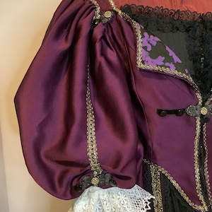 Może przedstawiać: Zbliżenie na odzież w stylu vintage z rękawem z głębokiego fioletowego satyny. Ubranie posiada złote wykończenia, czarną koronkę i ozdobne guziki. Biały koronkowa mankiet jest widoczny na końcu rękawa. Gorset jest czarny z fioletowymi kwiatowymi akcentami.