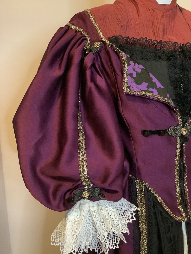 Może przedstawiać: Zbliżenie na odzież w stylu vintage z rękawem z głębokiego fioletowego satyny. Ubranie posiada złote wykończenia, czarną koronkę i ozdobne guziki. Biały koronkowa mankiet jest widoczny na końcu rękawa. Gorset jest czarny z fioletowymi kwiatowymi akcentami.