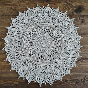 Könnte beinhalten: Ein cremefarbenes, handgefertigtes Häkel-Tischset mit einem detaillierten, kreisförmigen Design. Das Tischset weist ein zentrales Spiralmuster auf, das von konzentrischen Ringen aus komplizierter Spitze und einem gewellten Rand umgeben ist. Dieses dekorative Stück ist perfekt.
