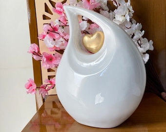 May include: White ceramic vase shaped like a teardrop with a gold heart cutout. The vase is sitting on a wooden surface.