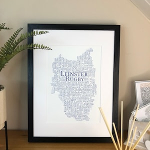May include: A white framed print with a blue word cloud shaped like the island of Ireland. The word cloud includes the names of towns and cities in Ireland, as well as the names of rugby teams. The text "LEINSTER RUGBY" is prominently displayed in the center of the word cloud.