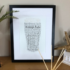 May include: A black and white word cloud art print featuring the names of pubs in Armagh, Northern Ireland. The print is framed in a black frame and is displayed on a wooden surface.