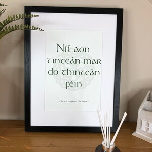 May include: A framed print with a black frame and a white background. The print features a Celtic knot design and the Irish phrase "Nil aon tinteán mar do thinteán féin" which translates to "There's no place like home".