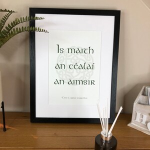 May include: A framed print with a black frame and a white background. The print features a green Celtic knot design with the Irish phrase "Is maith an cealáí an aimsir" in green text. The text below the design reads "Time is a great storyteller."