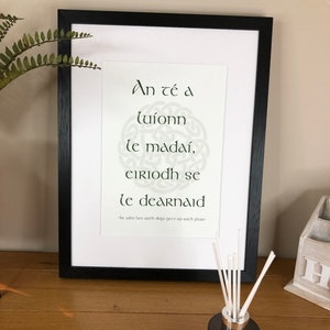 May include: A framed print with a white background and black frame. The print features a green Celtic knot design with the Irish Gaelic phrase "An té a luíonn le madaí, eiriodh se le dearnaid" which translates to "He who lies with dogs gets up with fleas".