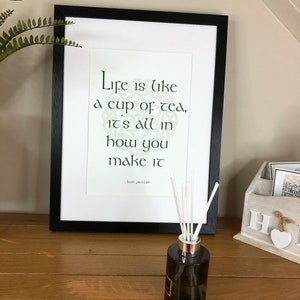 May include: A framed print with the quote "Life is like a cup of tea, it's all in how you make it" in green lettering. A dark brown glass diffuser bottle with white reeds sits in front of the frame. The frame is black.