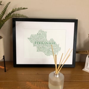 Puede incluir: Una impresión enmarcada de un mapa de nube de palabras de Fermanagh, Irlanda del Norte. El mapa está formado por texto verde, con el nombre "Fermanagh" en un tamaño de fuente más grande. La impresión está en un marco negro y está sobre una superficie de madera.