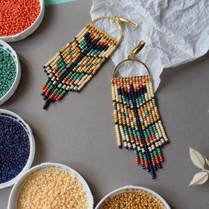 May include: A pair of long, beaded earrings with a geometric pattern in shades of cream, orange, blue, and green. The earrings are made with small seed beads and have a gold-toned hook closure.
