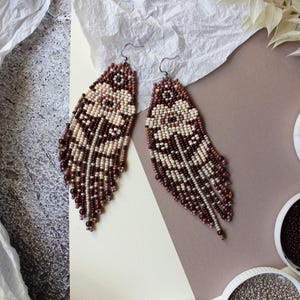 May include: Handmade beaded earrings in a feather design. The earrings are made with cream, burgundy, and brown beads. The earrings are displayed on a neutral background with decorative elements. The earrings are a unique accessory.