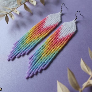 Op de afbeelding: Een paar lange, regenboogkleurige kralen oorbellen met witte uiteinden. De oorbellen zijn gemaakt van kleine, kleurrijke kralen en hangen aan zilveren haken.
