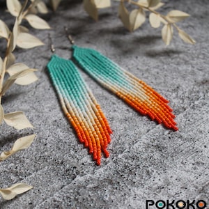 May include: Long beaded earrings in a gradient of colours. The earrings transition from turquoise to white, then to orange, and finally to red at the ends. The earrings are displayed on a textured grey surface.