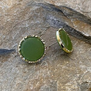 May include: A pair of green round stud earrings with a gold-toned border. The earrings are on a textured brown surface.