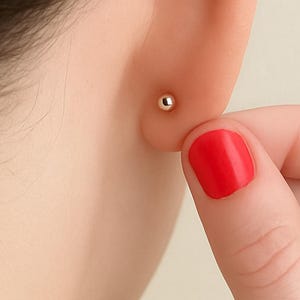 May include: A close-up of a person's ear with a small, round, gold stud earring. The person's hand, with red nail polish, is touching the ear. The background is a plain, light-colored wall.
