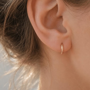 Puede incluir: Un pequeño pendiente de aro de oro pulido colocado en el lóbulo de la oreja. El pendiente presenta un diseño circular sencillo con una superficie metálica lisa y reflectante. Es una pieza de joyería minimalista sobre la oreja.