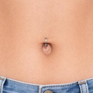 May include: Close-up of a belly button piercing. The piercing is a small, silver hoop. The image also shows the lower portion of a person's torso, including the top of blue denim jeans.