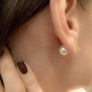 May include: A close-up of a person's ear wearing a pearl earring. The earring has a large, round, white pearl and a small, gold-colored stud. The person's skin is fair, and their hair is dark brown.