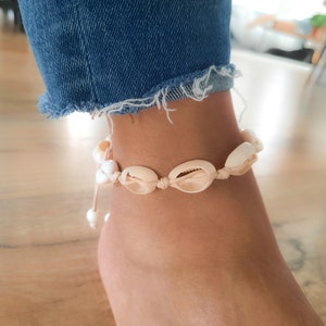 May include: A white and beige seashell anklet with a white string. The anklet is tied around a person's ankle.