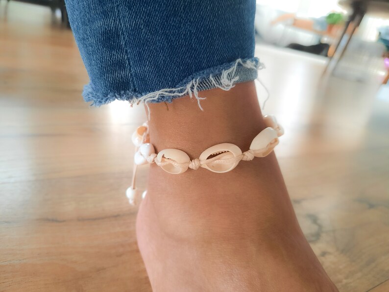 May include: A white and beige seashell anklet with a white string. The anklet is tied around a person's ankle.