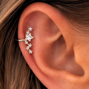 Könnte beinhalten: Ein silberner Ohrklemmen-Schmuck mit floralem Design und mehreren kleinen, klaren Edelsteinen. Der Schmuck verläuft entlang des Ohrknorpels, mit einer größeren Blume in der Mitte und kleineren sternförmigen Akzenten. Das Schmuckstück befindet sich am Ohr.