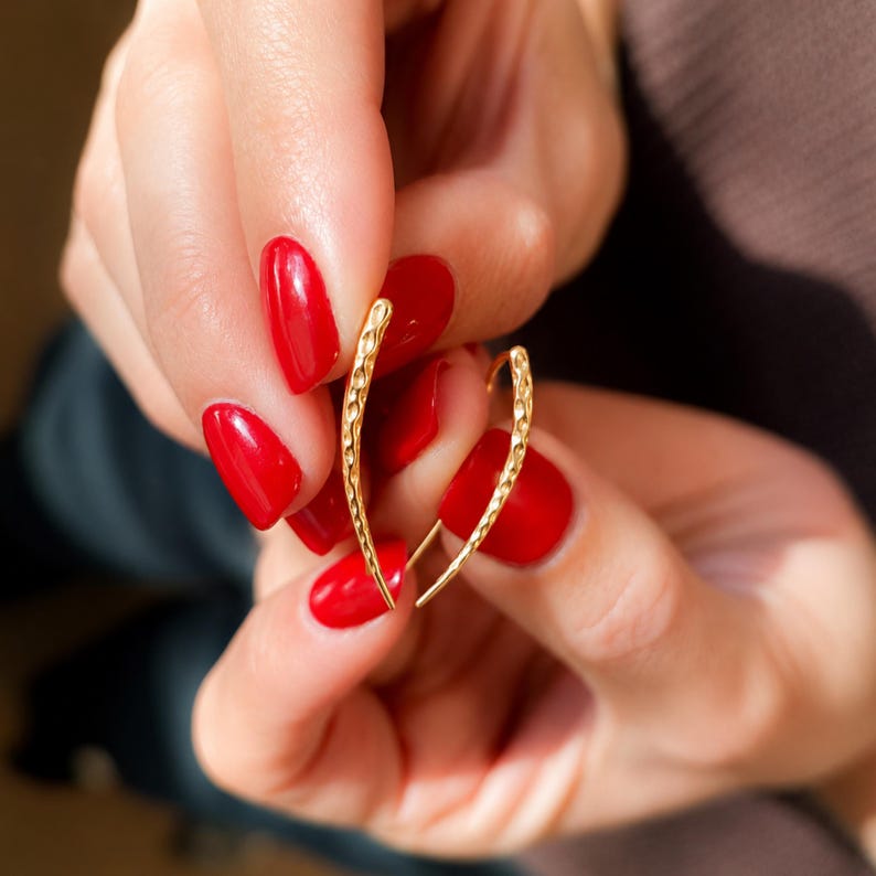 May include: A pair of gold-colored, textured earrings. The earrings have a curved, elongated design. The earrings are held between two hands with red nail polish.