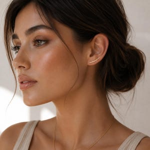 May include: A close-up shot of a woman wearing a gold ear climber earring. The earring curves along the ear. A gold necklace is also visible. The woman has dark hair pulled back and is wearing a neutral-colored top.