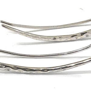 May include: A pair of silver-colored ear climbers. Each earring features a curved design with a textured, hammered finish on the main body and a smooth, curved hook for securing to the ear. The earrings are displayed on a white background.