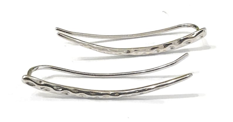May include: A pair of silver-colored ear climbers. Each earring features a curved design with a textured, hammered finish on the main body and a smooth, curved hook for securing to the ear. The earrings are displayed on a white background.