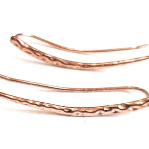May include: A pair of copper-colored ear climbers. The earrings have a hammered texture on the curved bar and a smooth, thin wire that curves up and around the ear. The earrings are displayed on a white background.