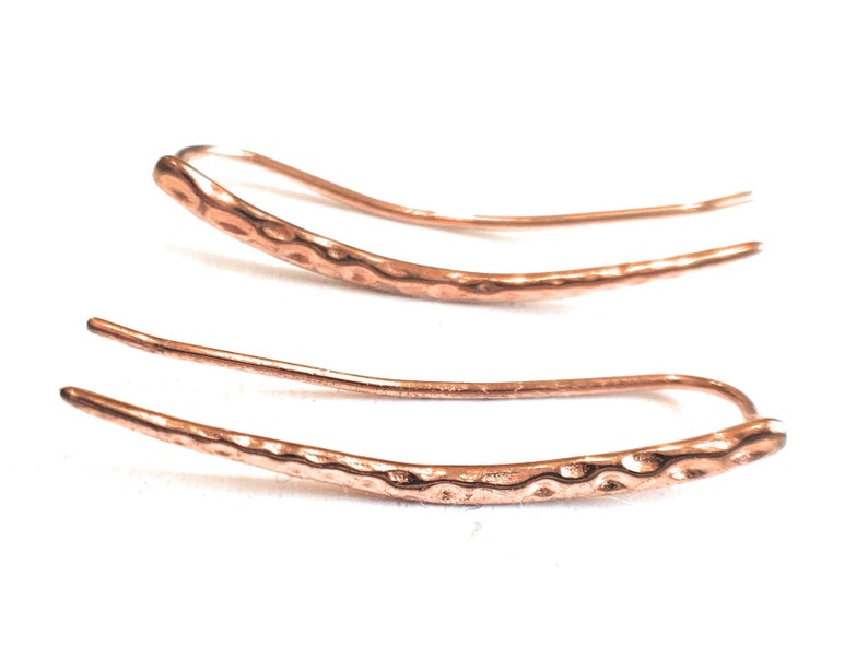 May include: A pair of copper-colored ear climbers. The earrings have a hammered texture on the curved bar and a smooth, thin wire that curves up and around the ear. The earrings are displayed on a white background.