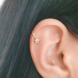 May include: A gold ear cuff with a flower-shaped charm set with clear gemstones. The cuff is worn on the ear, with the flower charm near the helix. The jewelry is set against a background of dark hair and skin.
